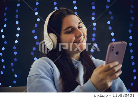 Young woman smiling with braces while listening favorite music and looking at phone against blurred blue background with lightening. Young woman smiling with braces while listening favorite music and looking at phone against blurred blue background with lightening. 110880401