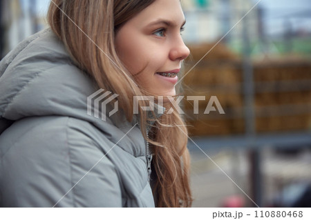 Side view portrait of youthful girl in winter coat smiling with braces, looking away against urban backdrop. Soft daylight. 110880468