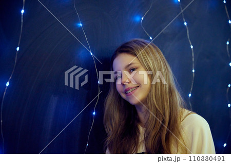 Close up photo of smiling teen girl with braces stands illuminated warm ambient lighting against blue background. Cozy indoor setting. Festive mood. 110880491