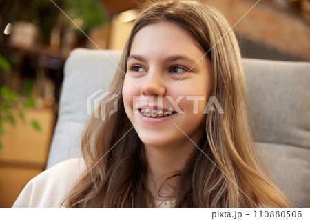 Close up portrait of young attractive girl smiling with teeth in braces sitting in arm chair and looking away. 110880506