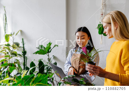 Happy middle age woman in yellow sweater in a flower shop with a purchased plant, talking with asian female seller in apron holding laptop surround plants in greenery.. 110882705