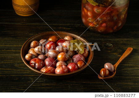 A bowl of pickled plums and a spoon on a rustic dark wooden table. A jar of plums in the background. Vegetarian healthy food concept 110883277