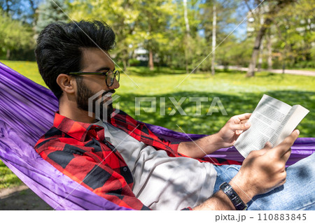 Contented young man in hammock reading a book Contented young man in hammock reading a book 110883845