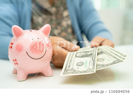 Asian elderly woman holding US dollar banknotes with pink piggy bank for saving money and insurance, poverty, financial problem in retirement. 110884056