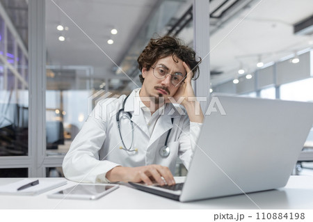 A man in a white medical coat with a stethoscope sits in front of a laptop computer in a medical office. Doctor thinking using computer 110884198
