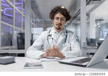 A doctor in a white coat and stethoscope sitting at a desk inside a medical office, using a laptop. Man serious looking camera, portrait 110884204