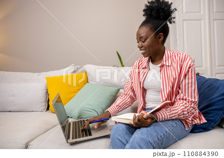 Woman with afro hairstyle writing notes in her panner and using laptop sitting on sofa. 110884390