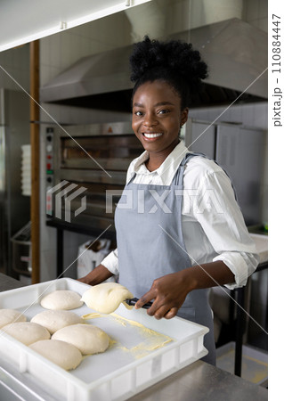 Smiling african american woman standing near the table with dough for pizza Smiling african american woman standing near the table with dough for pizza 110884447