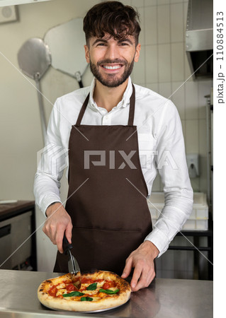 Smiling young cooker slicing pizza and looking contented 110884513
