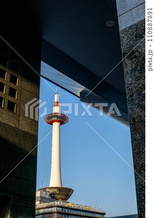 京都駅前の春、青空を背にそびえる京都タワーを駅ビルの壁を額縁に見る 110887891