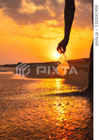Volunteer and plastic bottle, collecting waste on the sea beach, pollution and recycling concept. hand holds a transparent plastic bottle in water lying on a sandy beach during sunset or dawn 110892906