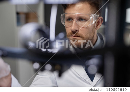 Medium close up shot of focused Caucasian male technician in protective goggles working in laboratory, look through blurred 3D printer components Medium close up shot of focused Caucasian male technician in protective goggles working in laboratory, look through blurred 3D printer components 110893610