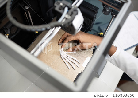 Top down view of unrecognizable technicians hand reaching for illuminated wrist bone model on metal hot bed in enclosed 3D printer 110893650