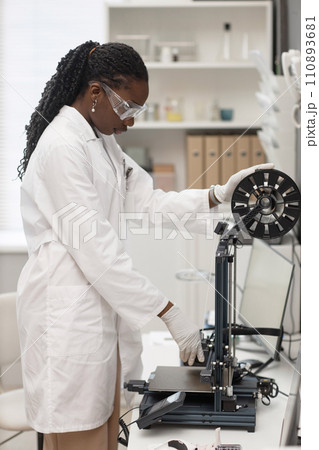 Vertical shot of Black woman engineer in lab coat and protective glasses setting up 3D printer for work in laboratory Vertical shot of Black woman engineer in lab coat and protective glasses setting up 3D printer for work in laboratory 110893681