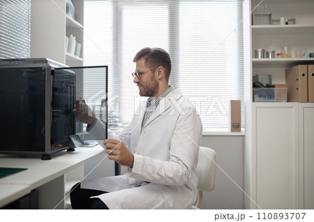 Smiling Caucasian male technician looking at 3D bone model in hand while opening enclosed 3D printer chamber and sitting on chair at table in laboratory 110893707