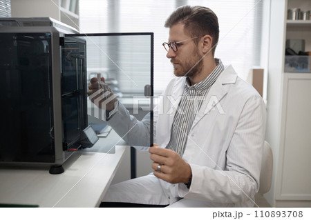 Medium long shot of Caucasian man engineer in lab coat and eyeglasses taking produced bone sample out of enclosed 3D printer in laboratory 110893708