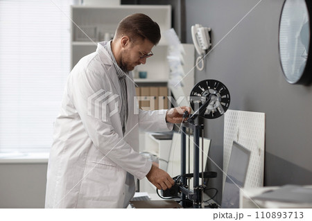 Caucasian male engineer in lab coat inserting white-colored filament strand into extruder of 3D printer in laboratory Caucasian male engineer in lab coat inserting white-colored filament strand into extruder of 3D printer in laboratory 110893713