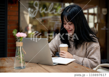A beautiful Asian woman is working remotely at a cafe in the city, working on her laptop. 110897617
