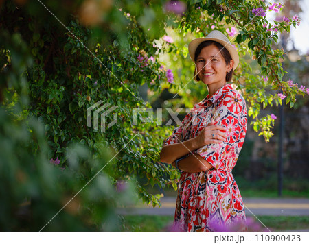 young asian woman stands in sunny green park, admiring blooming bougainvillea bush. The flower's bright colors are beautiful contrast to park's lush greenery. young asian woman stands in sunny green park, admiring blooming bougainvillea bush. The flower's bright colors are beautiful contrast to park's lush greenery. 110900423