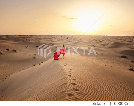 Desert adventure. Young arabian Woman in red silk dress in sands dunes of UAE desert at sunset, fantastic view. The Dubai Desert Conservation Reserve, United Arab Emirates. Desert adventure. Young arabian Woman in red silk dress in sands dunes of UAE desert at sunset, fantastic view. The Dubai Desert Conservation Reserve, United Arab Emirates. 110900859
