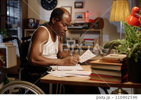 Black man in wheelchair writing information from paper into his notebook sitting at desk at home 110902596