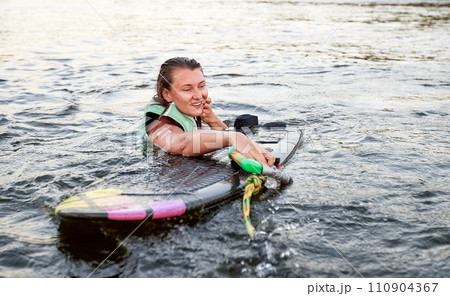 Young beautiful sporty girl in a green life jacket swims in the water with wakeboard and halyard in her hand. Happy sportswoman preparing for a steep wakeboard ride. Active lifestyle, healthy hobby. 110904367