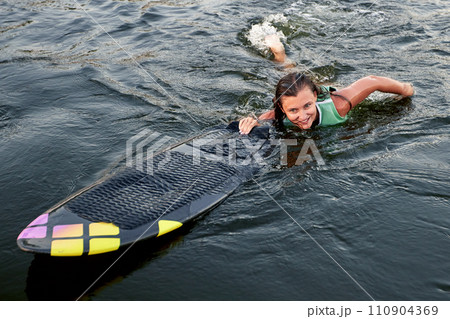Young beautiful sporty girl in a green life jacket swims in the water with a wakeboard in her hand. Happy sportswoman is preparing for a steep wakeboard ride. Active lifestyle, healthy hobby. Young beautiful sporty girl in a green life jacket swims in the water with a wakeboard in her hand. Happy sportswoman is preparing for a steep wakeboard ride. Active lifestyle, healthy hobby. 110904369