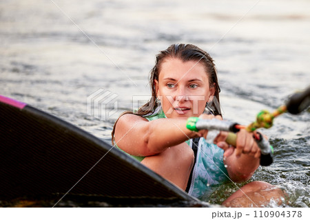 Young beautiful sporty girl in a green life jacket sitting in the water on a wakeboard with a halyard in her hand. Sportswoman is preparing for a steep wakeboard ride. Active lifestyle, healthy hobby. Young beautiful sporty girl in a green life jacket sitting in the water on a wakeboard with a halyard in her hand. Sportswoman is preparing for a steep wakeboard ride. Active lifestyle, healthy hobby. 110904378