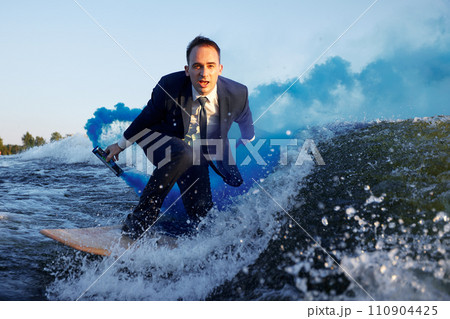 Young man in classic suit with blue smoke bomb in his hand rides wakeboard on a lake near city. Happy clerk escaped from stuffy office to take up active sport. Best summer leisure after routine work. Young man in classic suit with blue smoke bomb in his hand rides wakeboard on a lake near city. Happy clerk escaped from stuffy office to take up active sport. Best summer leisure after routine work. 110904425