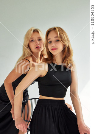 Half-length studio portrait of two young sexy women dressed in black clothes on white background. Stylish blonde girls posing with bar stool. Glamour models with languid look. 110904555