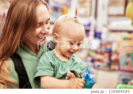 Young attractive mother with cute baby in her hands walking around toy store. Beautiful mom and her little blonde daughter are shopping in the mall and having fun. Family weekend, happy childhood. 110904595