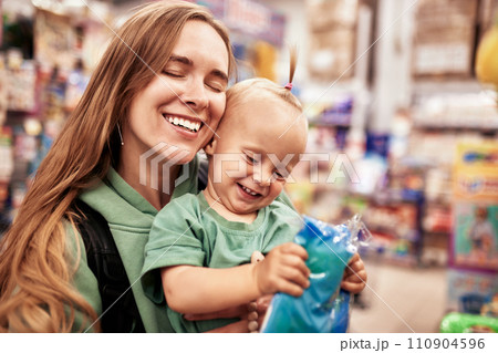 Young attractive mother with cute baby in her hands walking around toy store. Beautiful mom and her little blonde daughter are shopping in the mall and having fun. Family weekend, happy childhood. 110904596