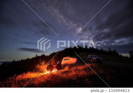 Happy couple staring at starry sky with clouds. Young people sitting in chairs and warming by campfire near tourist tent and off-road car. Travelling couple having a rest together. Night camping. Happy couple staring at starry sky with clouds. Young people sitting in chairs and warming by campfire near tourist tent and off-road car. Travelling couple having a rest together. Night camping. 110904623