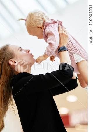 Young attractive mother lifts up her baby on outstretched arms in public place, shopping mall. Mom and little daughter in pink clothes play, relax and have fun while shopping. Family weekend in mall. 110904631