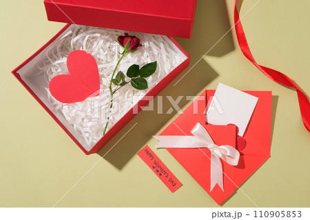 A gift box in red color containing a lot of kraft paper with a red paper heart and a branch of rose placed on. Red envelope with a ribbon bow displayed with a small paper of wishes A gift box in red color containing a lot of kraft paper with a red paper heart and a branch of rose placed on. Red envelope with a ribbon bow displayed with a small paper of wishes 110905853