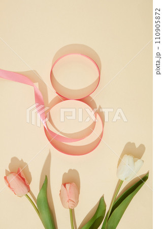 A ribbon in pink color arranged in a figure eight over beige background with few tulip flowers. Elegant card for International Women Day. Banner or leaflet for March 8 110905872