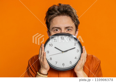 Serious strict young man holding office clock hiding behind checking time on watch obscuring face Serious strict young man holding office clock hiding behind checking time on watch obscuring face 110906378