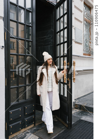 Young woman in stylish clothes walks out of a building with a large black door 110906475