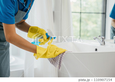 A maid wearing gloves diligently cleans bathroom sink and faucet eliminating limescale with spray detergent. Her focus on housework ensures purity hygiene and shining fixtures. spray cleaner 110907154