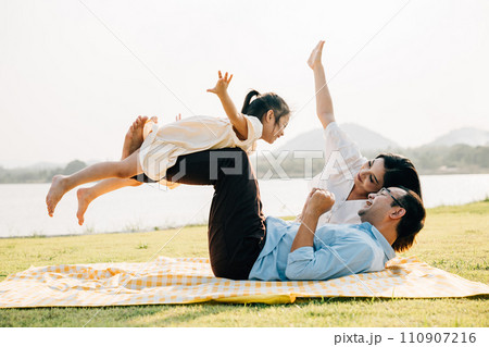 A family enjoys playful moments together. Father holds daughter high in air as she giggles and spreads her arms like wings, while mother watches with a smile outdoor. a precious moment of family fun A family enjoys playful moments together. Father holds daughter high in air as she giggles and spreads her arms like wings, while mother watches with a smile outdoor. a precious moment of family fun 110907216