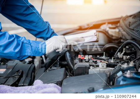 Skilled Asian mechanic in blue workwear jotting down notes on a paper while repairing a car in an auto repair shop. Close-up shot of hand. 110907244