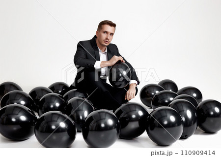 elegant caucasian man in suit tuxedo sitting on studio floor with a lot of black air balloons elegant caucasian man in suit tuxedo sitting on studio floor with a lot of black air balloons 110909414