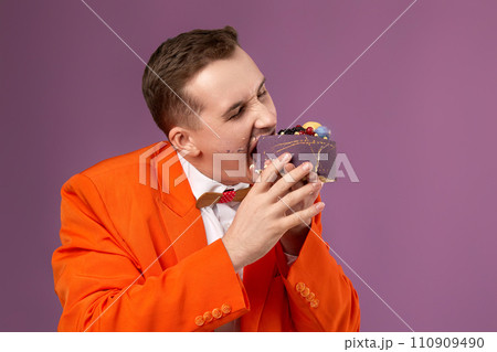 birthday man in orange jacket bites cake on purple background. copy space 110909490