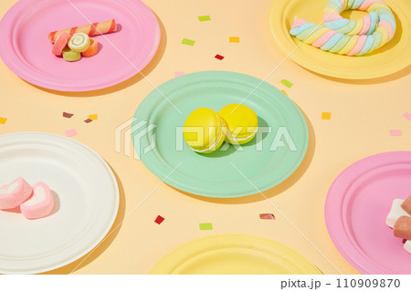 Cakes and marshmallows are displayed on colorful round plates. Confetti spread on pastel background. Copy scene for an advertisement with a birthday party theme. 110909870