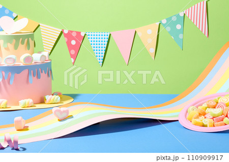 A string of birthday decorations on a green background. A birthday cake made from colored paper, a strip of colorful paper and a plate of gummy candy on a blue surface. Front view. 110909917