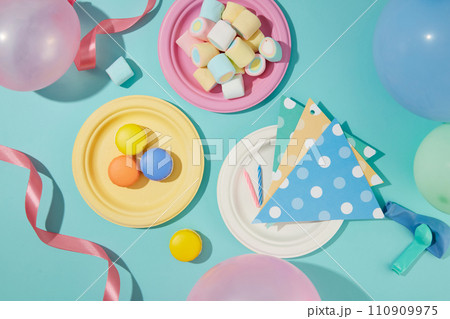 Flat lay of macarons, marshmallows and decorations displayed on round plates. Balloons and ribbons are decorated on a blue background. Attractive space. 110909975