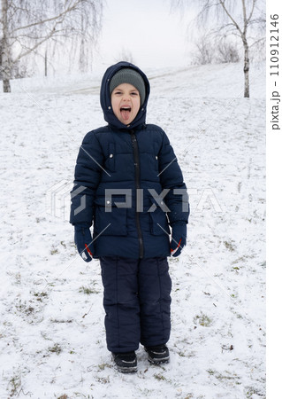 Funny little kid in warm overalls sticking out his tongue and grimacing in winter park. Funny little kid in warm overalls sticking out his tongue and grimacing in winter park. 110912146