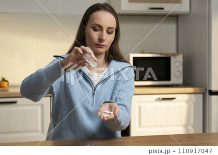 Woman pours pills into the palm of her hand while taking medicine sitting at the kitchen table 110917047