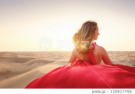 Desert adventure. Young arabian Woman in red silk dress in sands dunes of UAE desert at sunset, fantastic view. The Dubai Desert Conservation Reserve, United Arab Emirates. 110917708