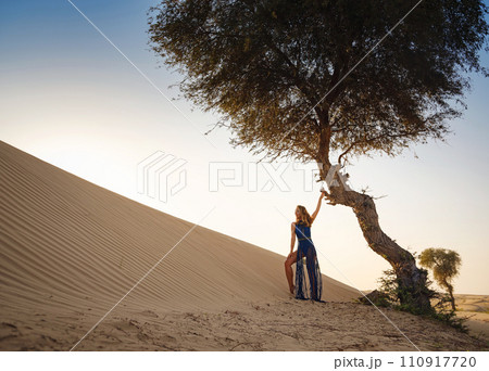 Desert adventure. Young arabian Woman in sexy boho blue makrame dress in sands dunes of UAE desert at sunset. The Dubai Desert Conservation Reserve, United Arab Emirates. 110917720